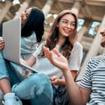 A group of students with laptops sit on the steps near the campus and communicate.
